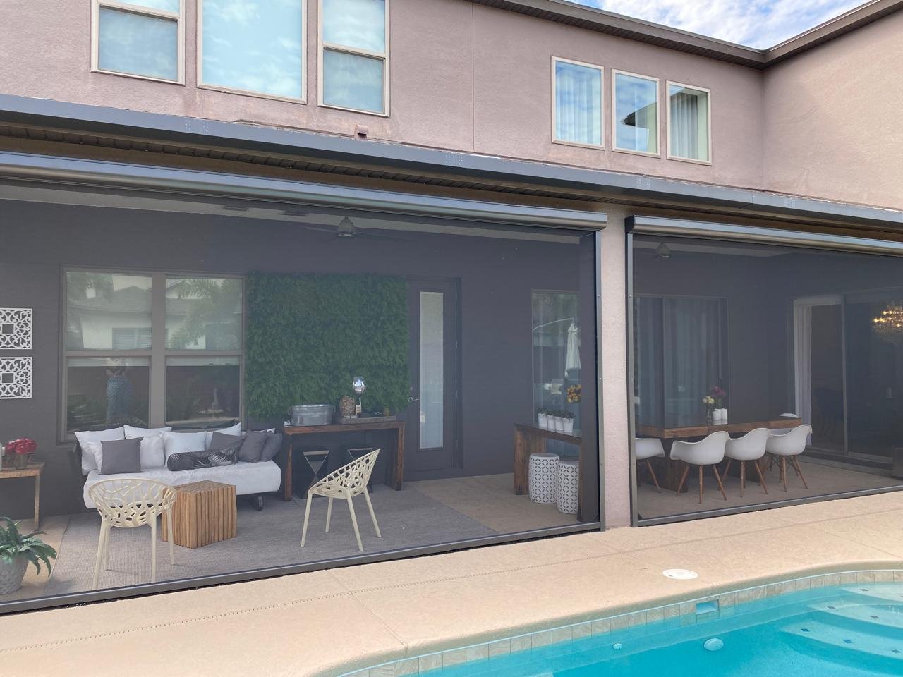 Covered patio lounge and dining area behind lowered outdoor shade screens, beside a pool
