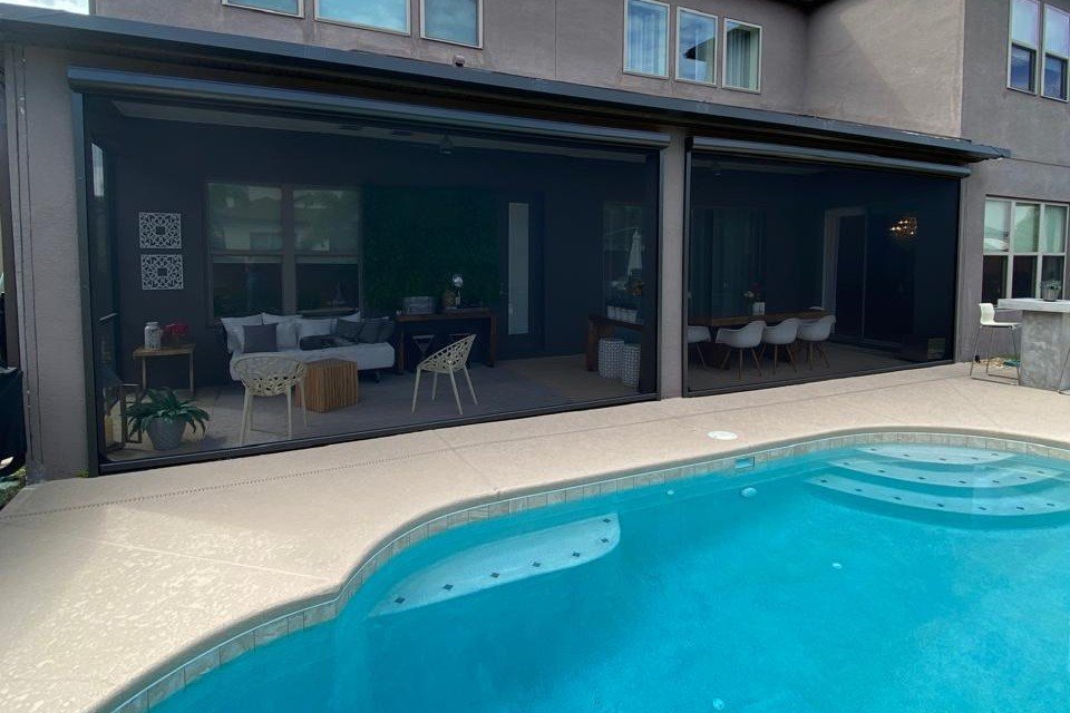 Covered patio lounge and dining area behind lowered outdoor shade screens, next to a pool